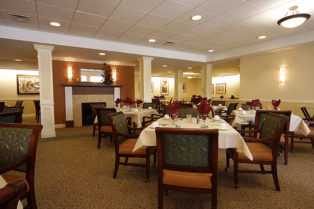 A dining room with multiple tables covered in white tablecloths and set with silverware and folded red napkins. The room features green upholstered chairs with wooden frames, a carpeted floor, a fireplace with decorative items on the mantel, and framed artwork on the walls. The ceiling has recessed lighting and a hanging light fixture.
