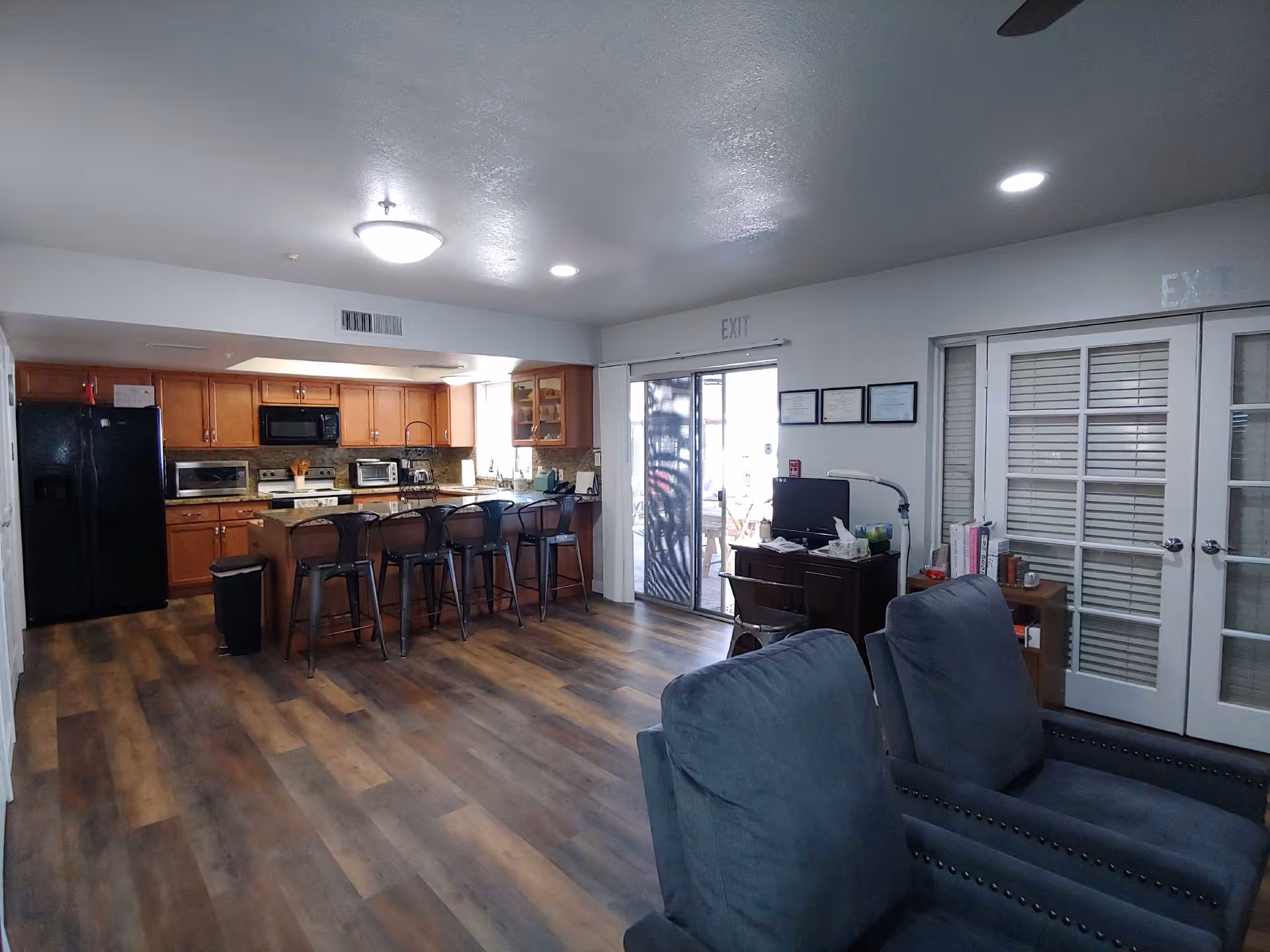 Interior view of a senior living facility showing a kitchen area with wooden cabinets, black refrigerator, microwave, toaster ovens, and a kitchen island with five black bar stools. Adjacent to the kitchen is a small office desk with a computer and chair, and two gray upholstered armchairs in the foreground. The room has wood-style flooring and white walls with glass-paneled double doors and a sliding glass door marked with exit signs.