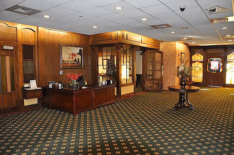 Interior view of a spacious lobby area with patterned carpet, wooden reception desk with flowers and coffee setup, wood-paneled walls, decorative stained glass windows, and a round table with a floral arrangement.