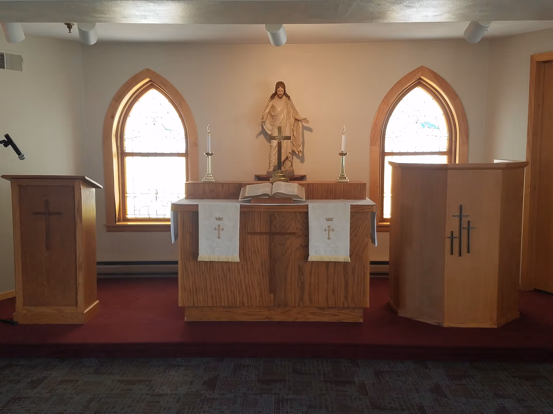 Interior of a small chapel with a wooden altar in the center, two wooden podiums on either side, two arched stained glass windows behind the altar, a statue of Jesus above the altar, two candles, and an open Bible on the altar.
