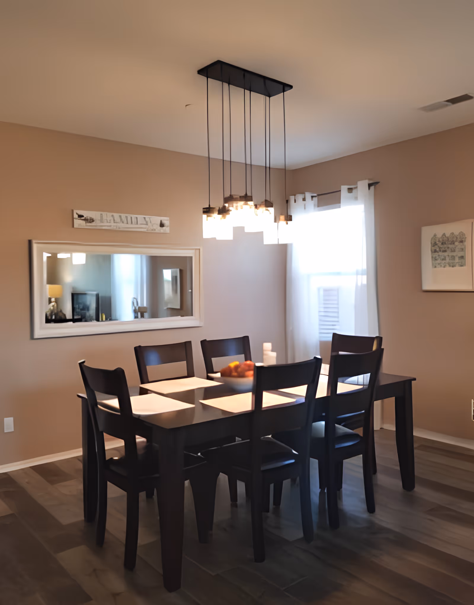 A dining room featuring a dark wood table with six chairs, place settings, a bowl of fruit, a pendant chandelier, and a wall mirror.