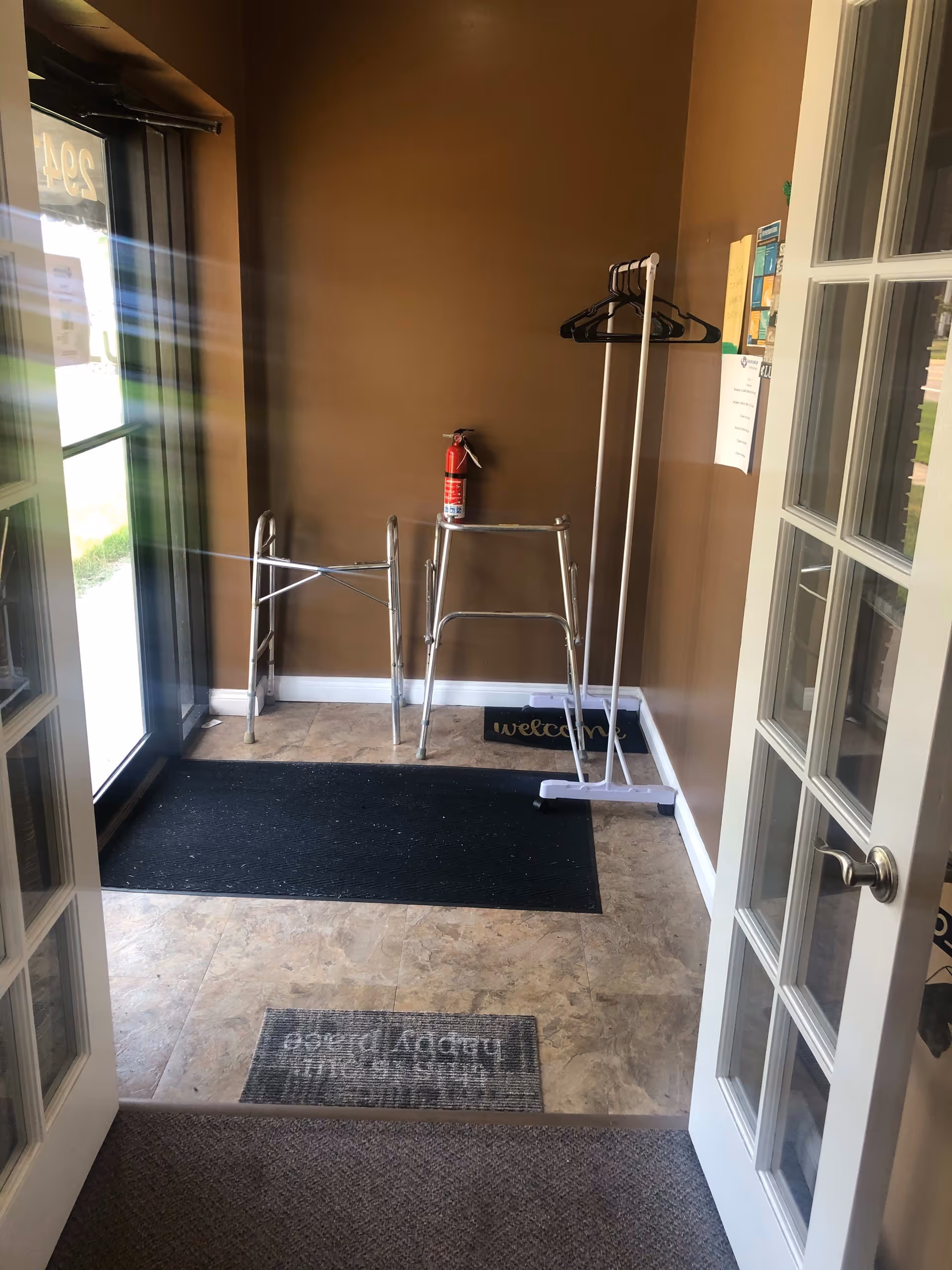 Entrance area of a facility with a glass door open, showing a tiled floor with two black mats and a welcome mat. There are two walkers and a white coat rack with hangers against a brown wall. A fire extinguisher is mounted on the wall above the walkers.