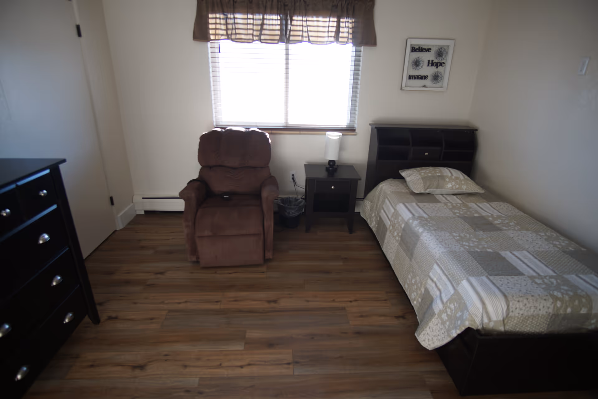 A small bedroom with a single bed covered in a beige and gray patchwork quilt, a brown recliner chair, a black nightstand with a lamp, a black dresser with silver knobs, and a window with blinds and a brown valance.
