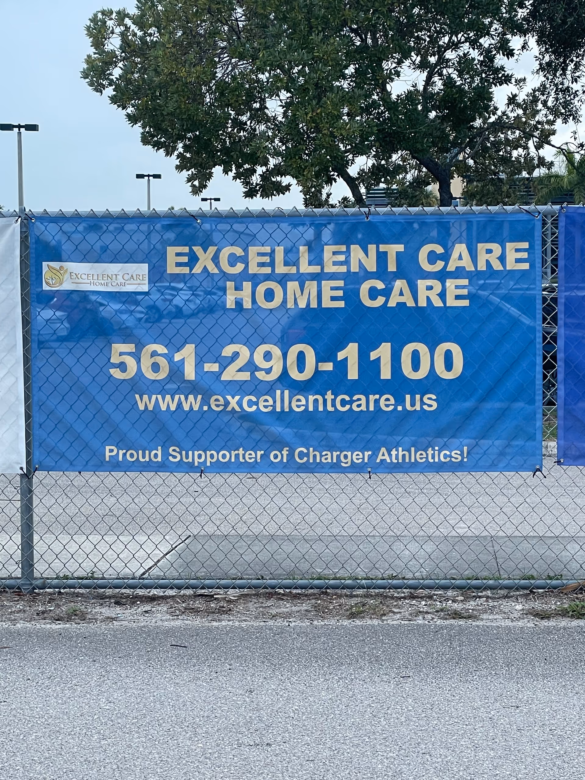 Blue banner on a chain-link fence advertising Excellent Care Home Care with phone and website.