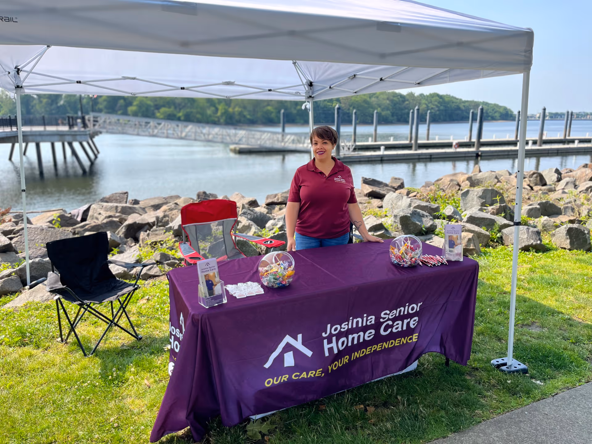 A staff member stands beside a purple Josinia Senior Home Care booth outdoors by the water.