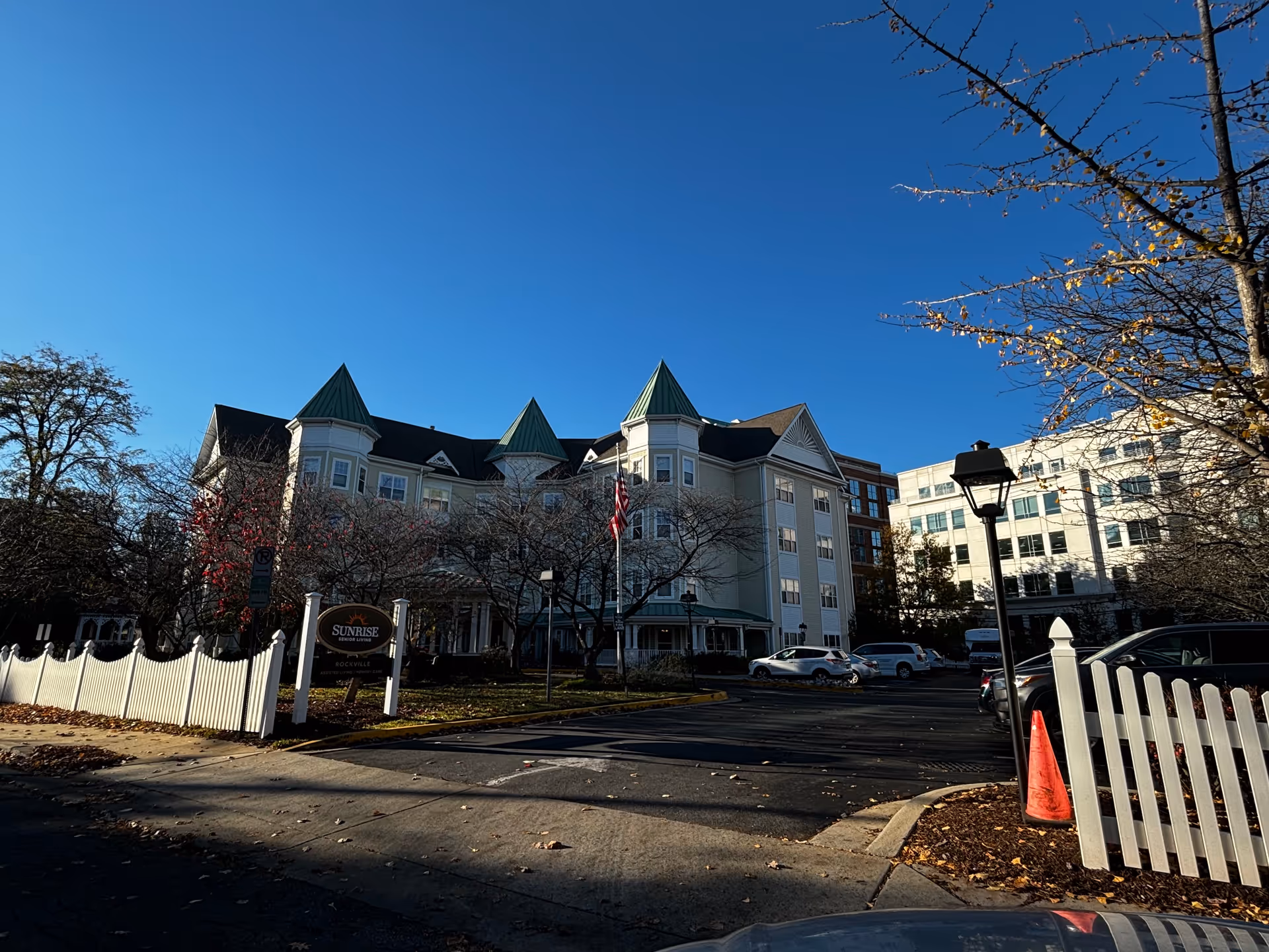Front exterior of the Sunrise of Rockville senior living building with green turrets, a white picket fence, driveway and parked cars under a clear blue sky.