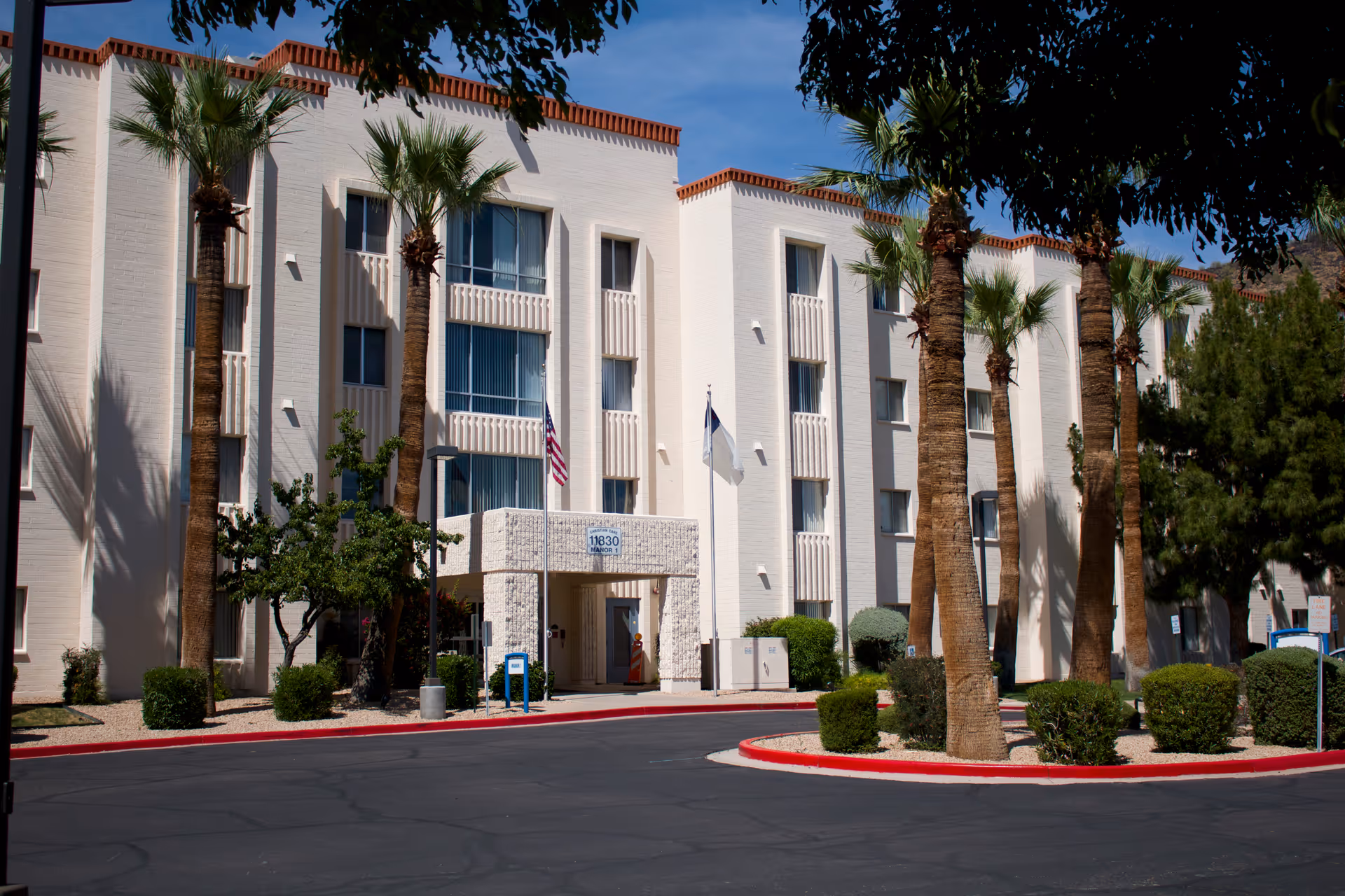 Exterior view of a three-story white building with palm trees and bushes in front, featuring an entrance with a covered walkway and two flags, one American and one white, under a clear blue sky.