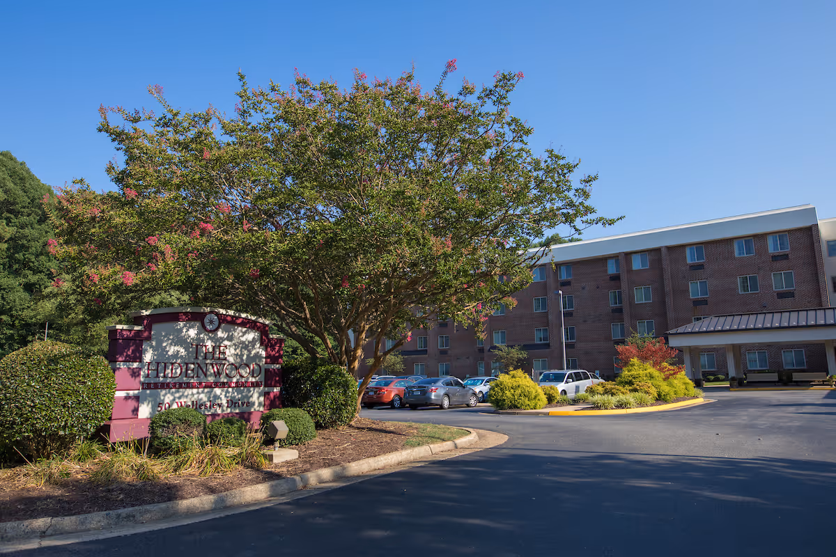 Front entrance of The Hidenwood Retirement Community showing the building, landscaped sign, large tree, and parked cars under a clear blue sky.