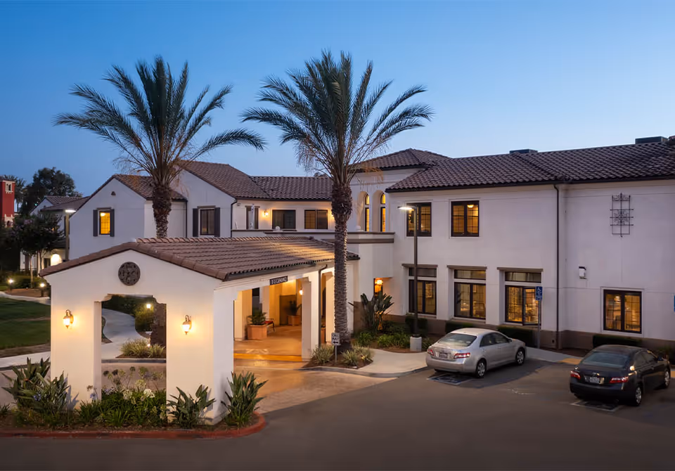 Two-story white senior living building with a shaded entrance, palm trees, and parked cars.