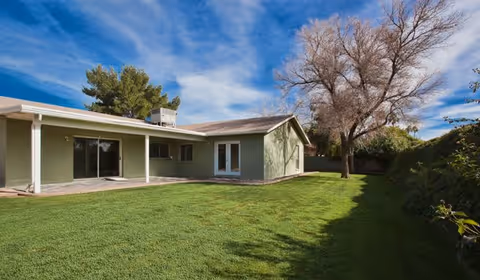 Exterior view of a single-story green building with a covered patio area, surrounded by a well-maintained lawn and trees under a blue sky with some clouds.