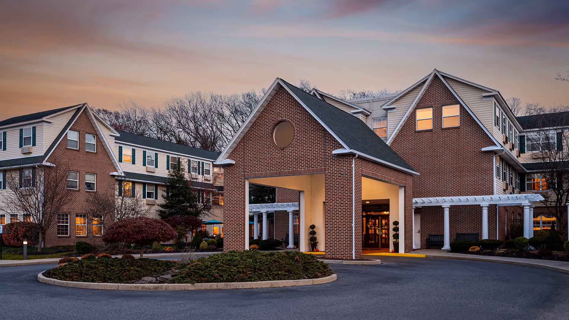Exterior view of a senior living facility building at dusk with a covered entrance, brick facade, multiple windows with lights on, and landscaped roundabout with shrubs and trees.