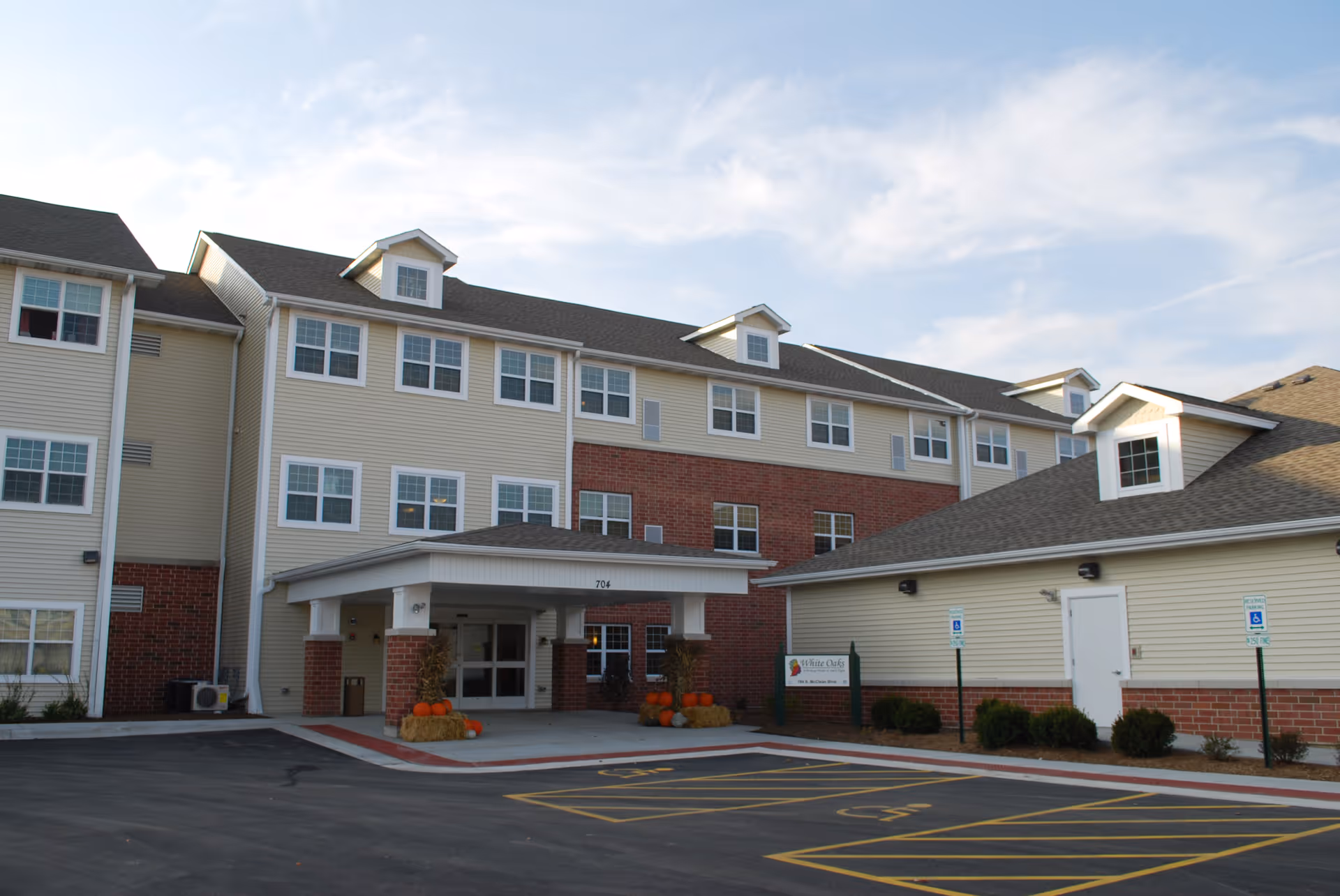 Front entrance of a three-story senior living building with beige siding and brick accents, a covered drop-off, and pumpkins decorating the doorway.