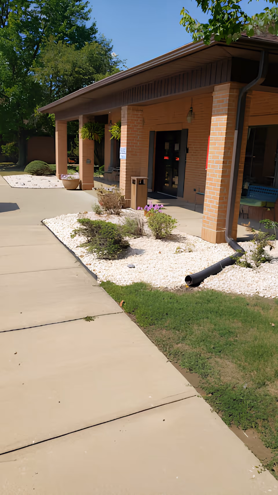 Covered brick entrance of a single-story health center with a sidewalk, landscaped white-rock beds, and potted plants.