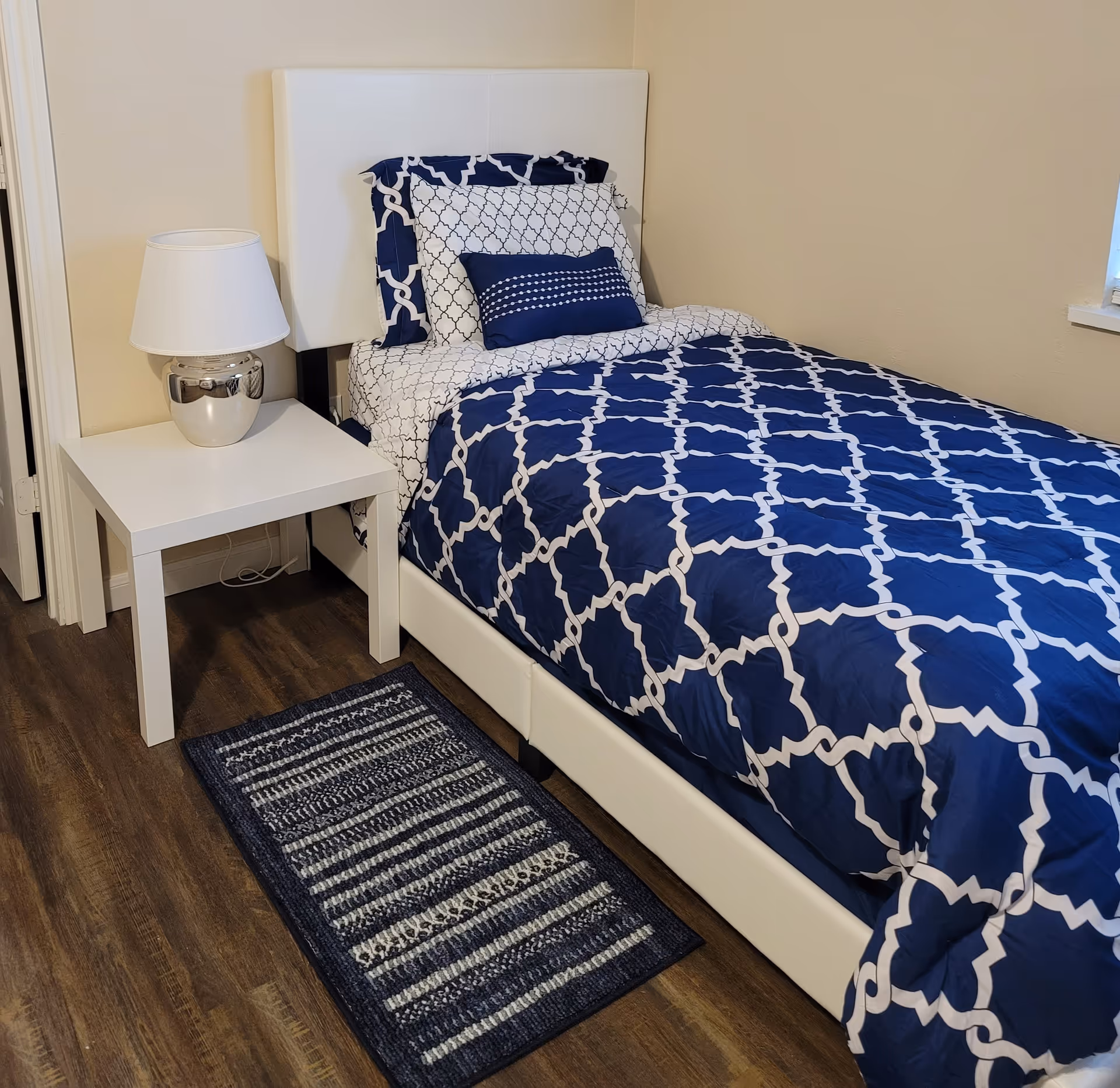 A neatly made single bed with a white upholstered headboard and blue and white patterned bedding. Next to the bed is a white nightstand with a silver and white lamp on it. The floor is wooden with a small blue and white striped rug beside the bed. The walls are beige and there is a window partially visible on the right side.