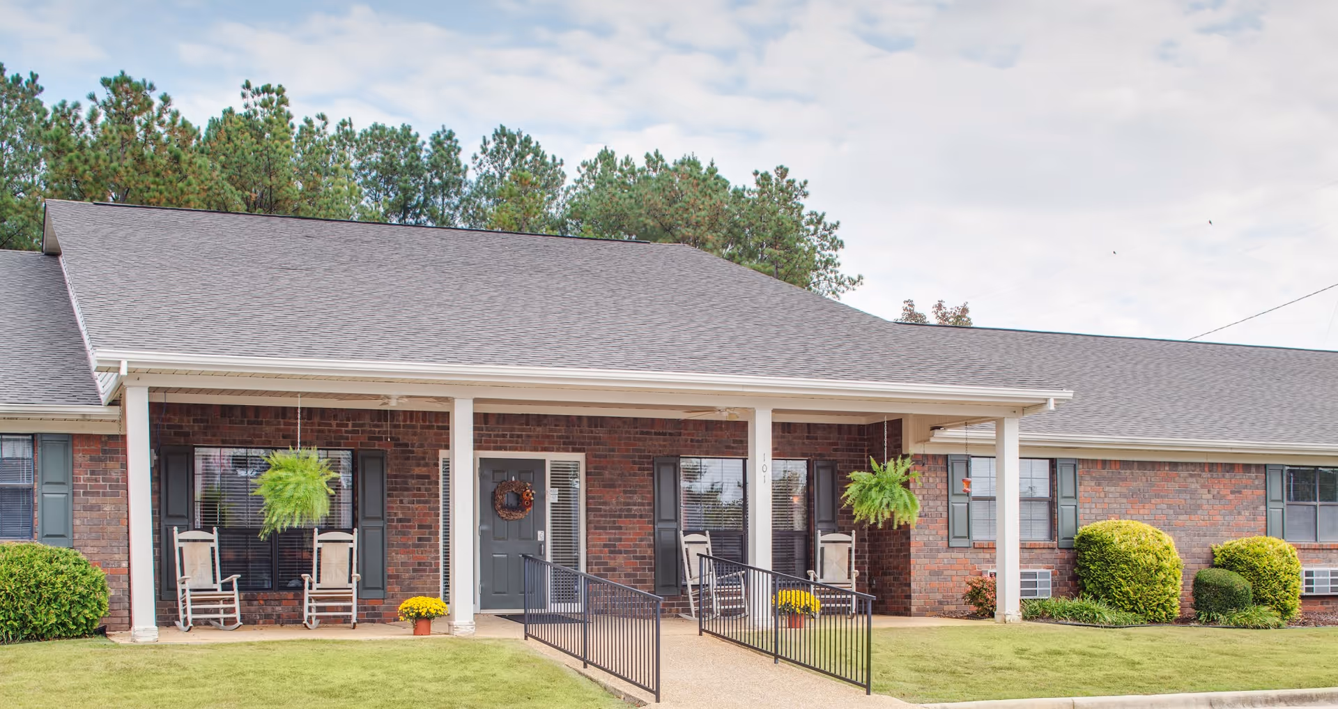 Single-story brick senior living facility with a front porch, rocking chairs, and hanging ferns.