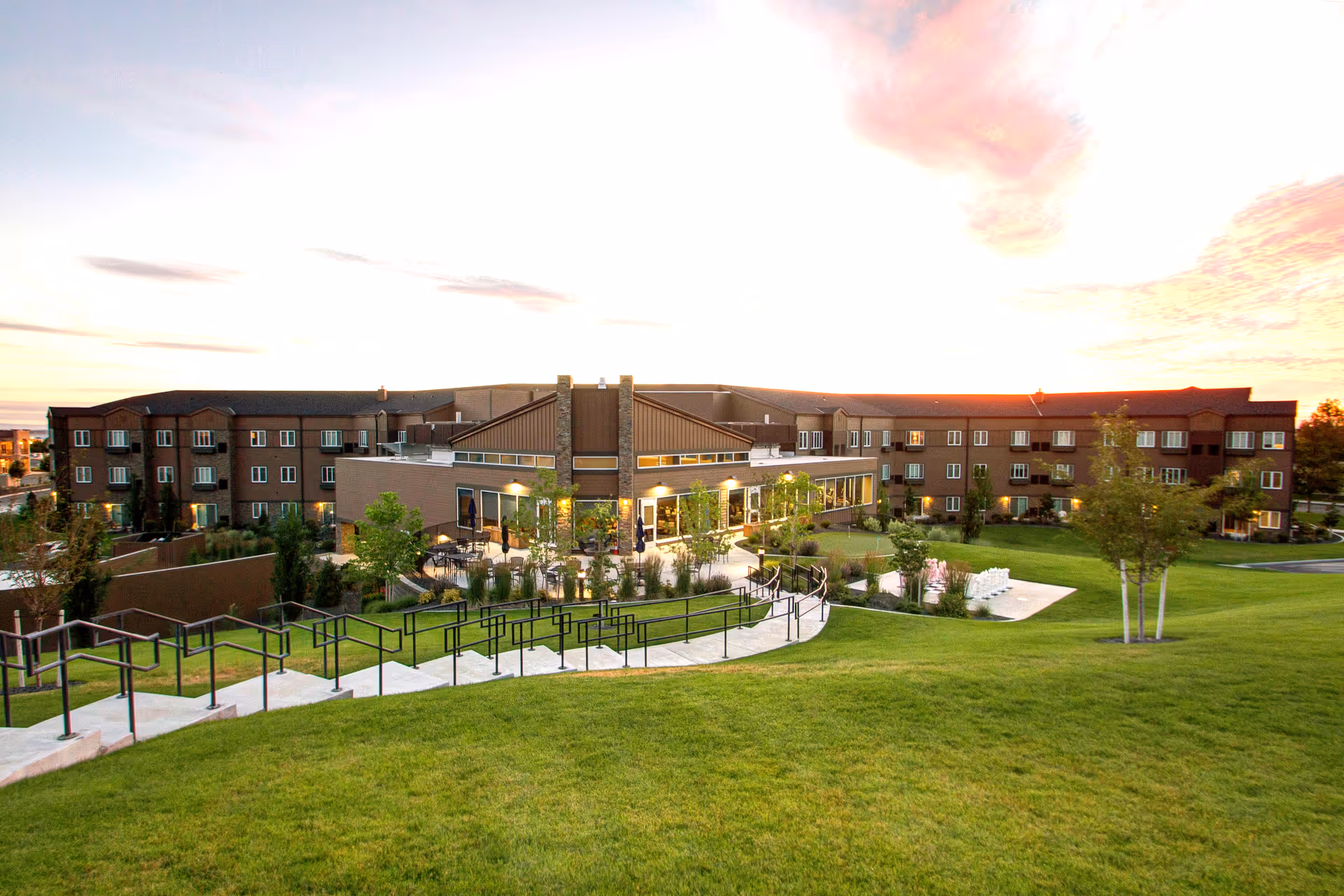 Wide exterior view of a large senior living facility building named Ciel of Tri-Cities during sunset, with a well-maintained green lawn, trees, and a paved walkway with handrails leading to the entrance area.