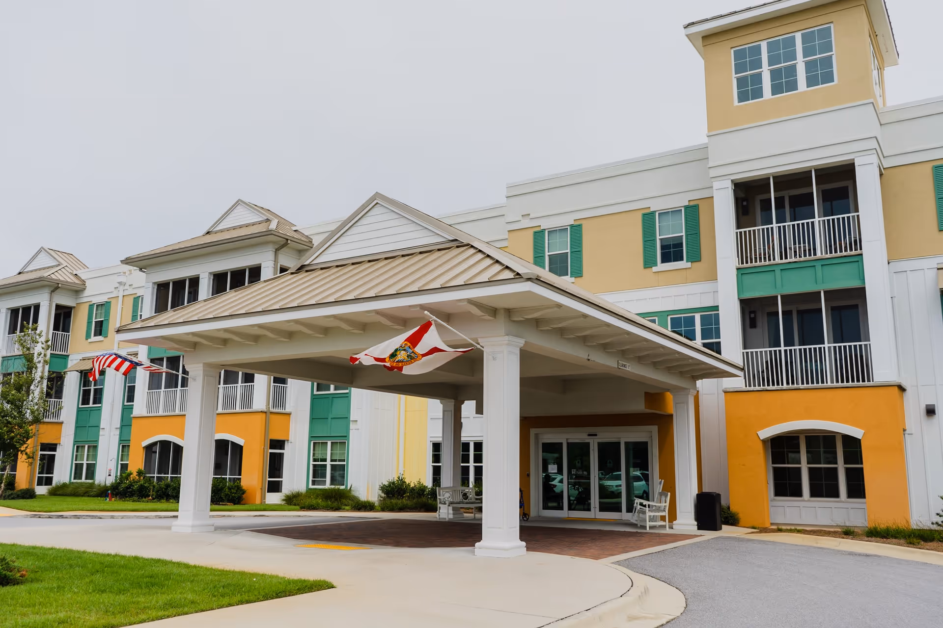 Exterior view of The Arbors of Gulf Breeze senior living facility showing the main entrance with a covered drop-off area, multiple windows, balconies, and colorful yellow, green, and white building facade with flags flying near the entrance.