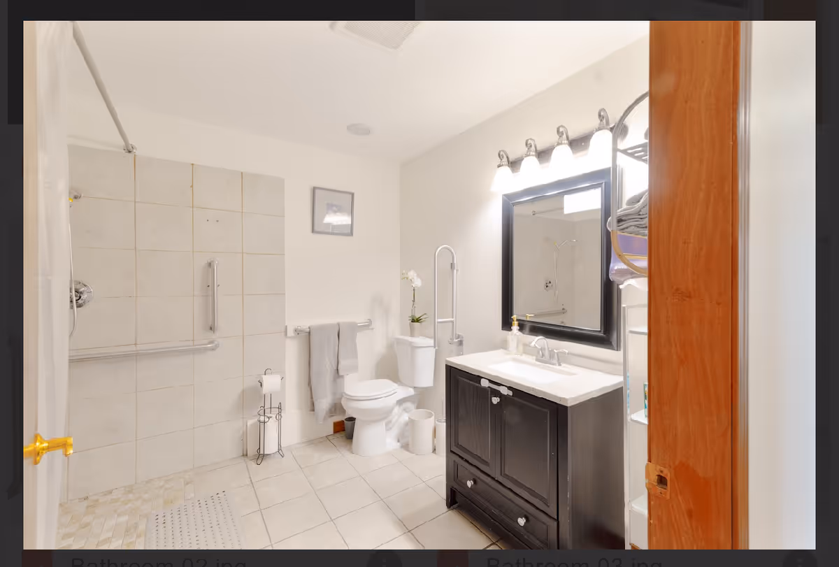 Bright bathroom with tiled shower, toilet, and dark vanity beneath a large mirror.