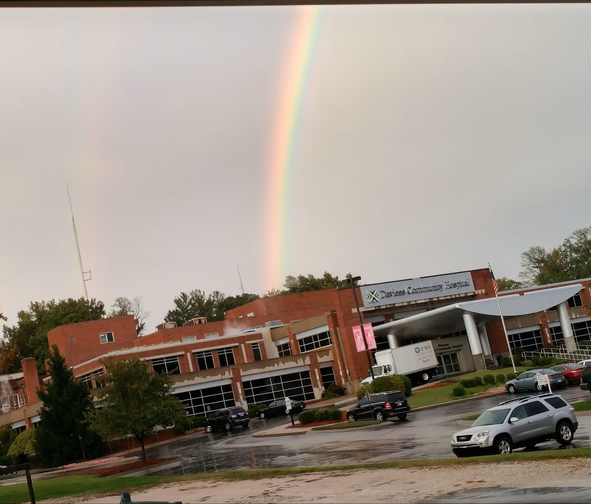 Wide view of a brick hospital building with a rainbow across a cloudy sky.