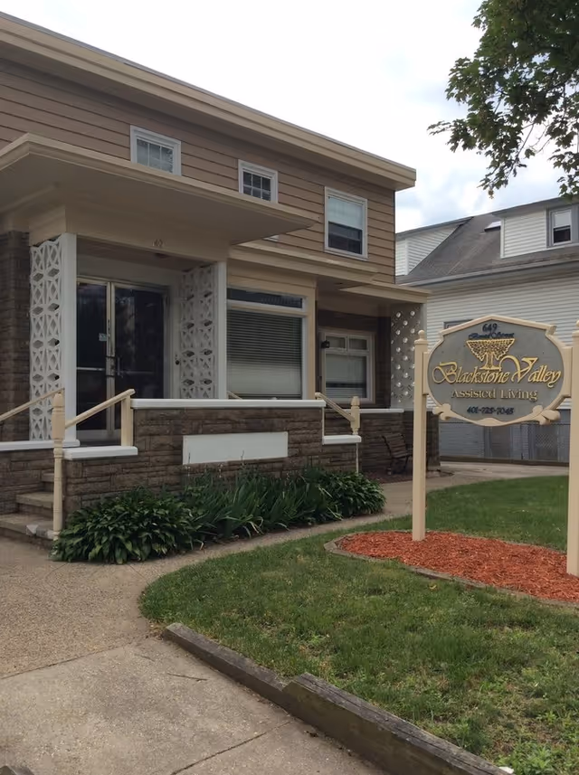 Exterior view of Blackstone Valley Assisted Living facility showing the front entrance with steps, a small garden with green plants, and a sign displaying the facility name and phone number.