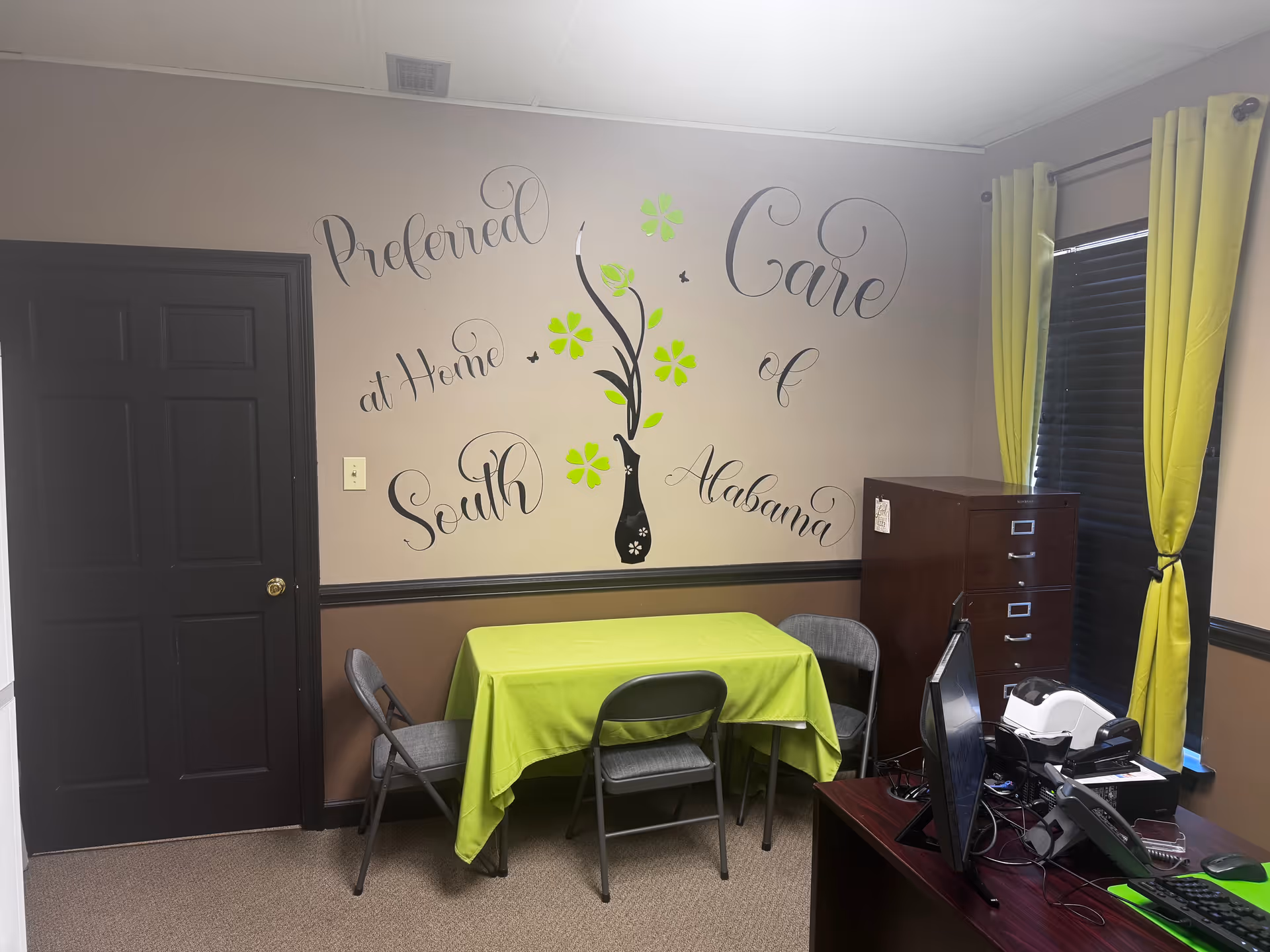Small interior activity room with lime-green tablecloth, chairs, desk and computer, and wall mural.
