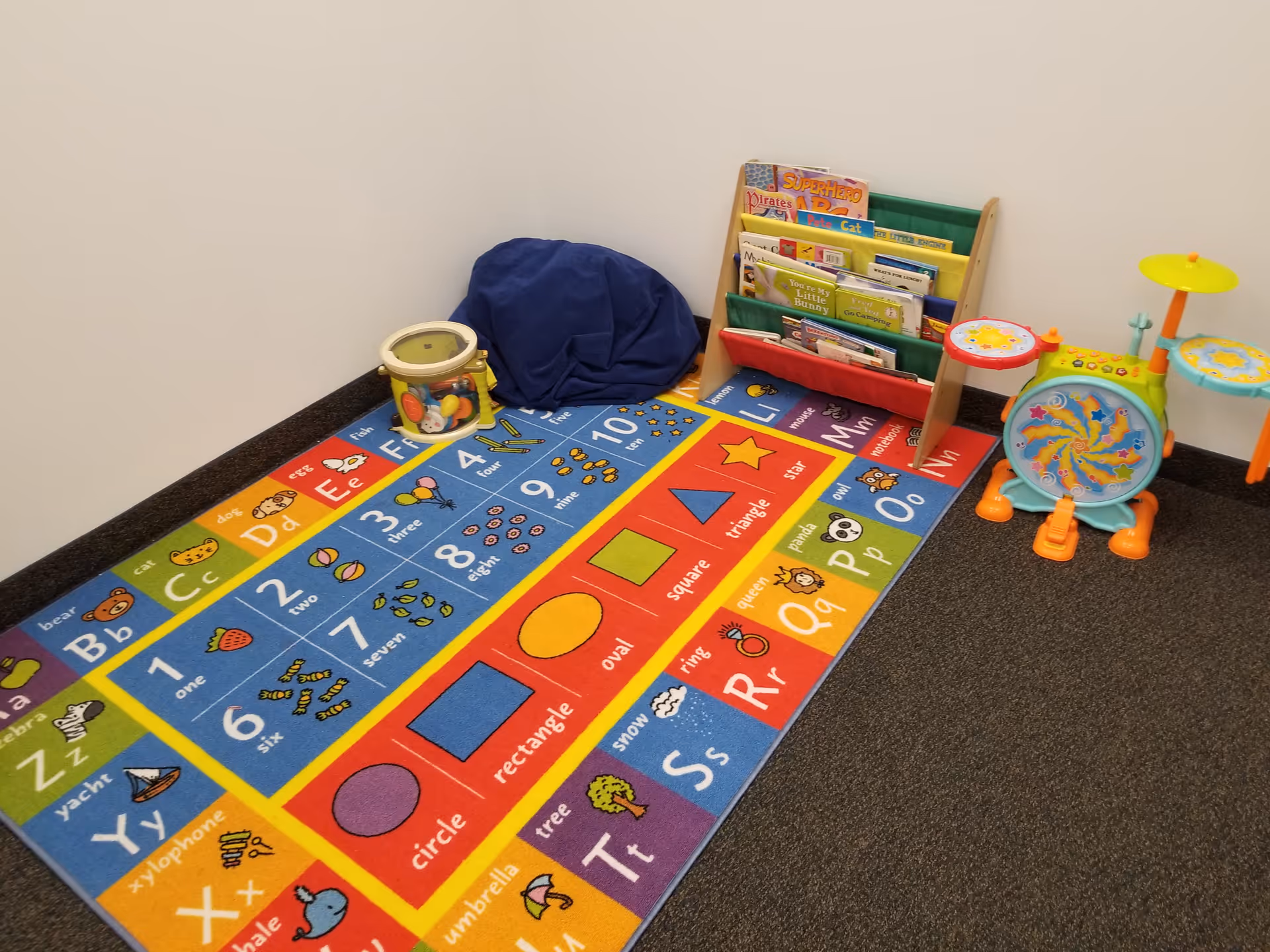 Colorful children's play area with alphabet rug, bookshelf, bean bag, and drums in corner.