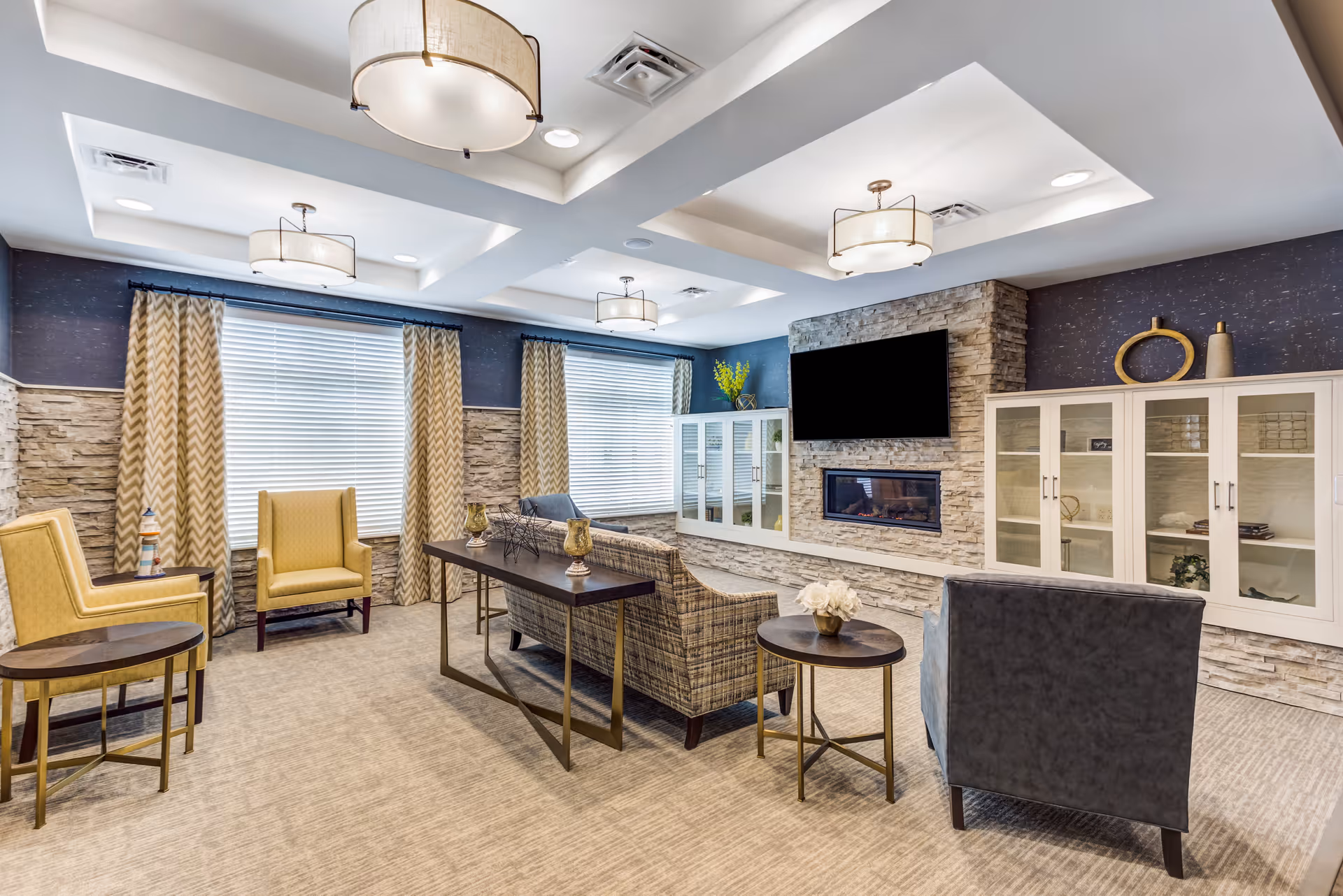 A bright and modern senior living facility common area with a stone accent wall featuring a mounted flat-screen TV above a built-in electric fireplace. The room has large windows with chevron-patterned curtains, a mix of yellow and gray armchairs, a plaid sofa, and several small tables with decorative items. The ceiling has recessed lighting and stylish drum pendant lights.