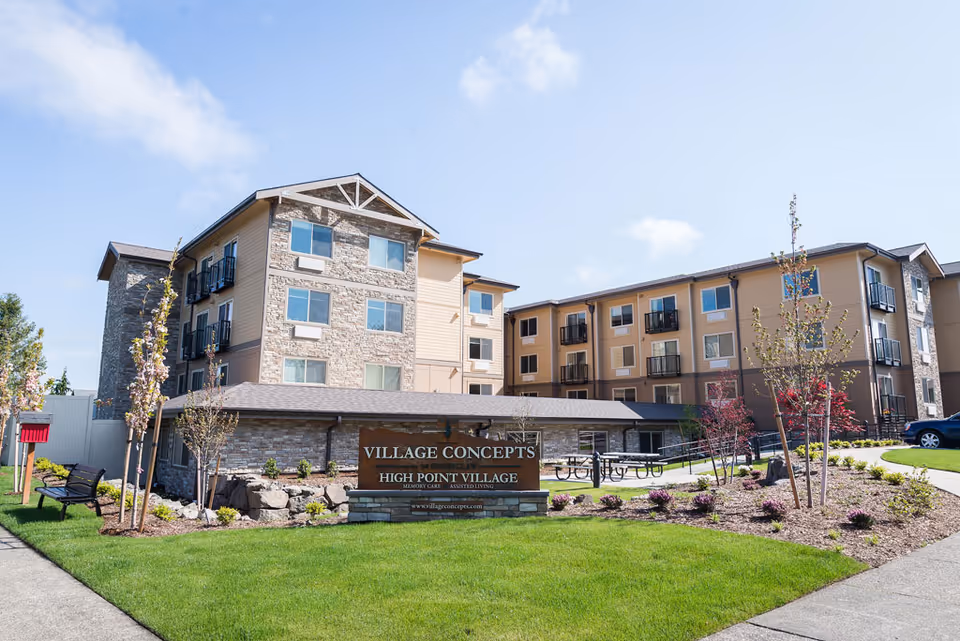 Exterior front of a multi-story senior living building with landscaped lawn and a sign reading "Village Concepts High Point Village".
