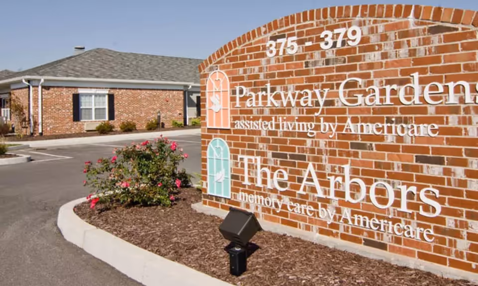 Brick entrance sign reading 'Parkway Gardens' and 'The Arbors' with landscaping and a low brick building in the background.