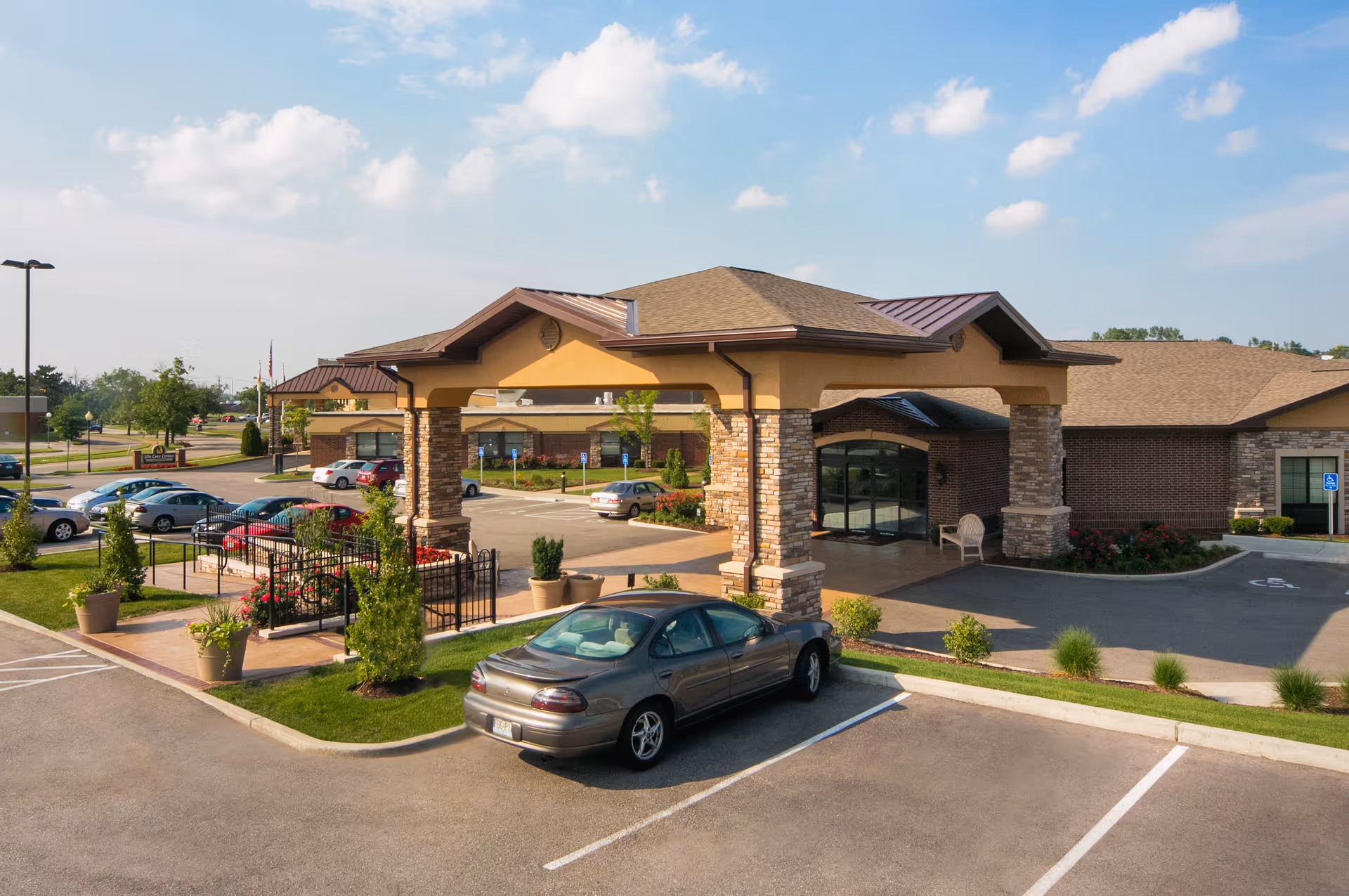 The front entrance of a senior living facility with a covered porte-cochere, landscaped planters, and parked cars in the parking lot.