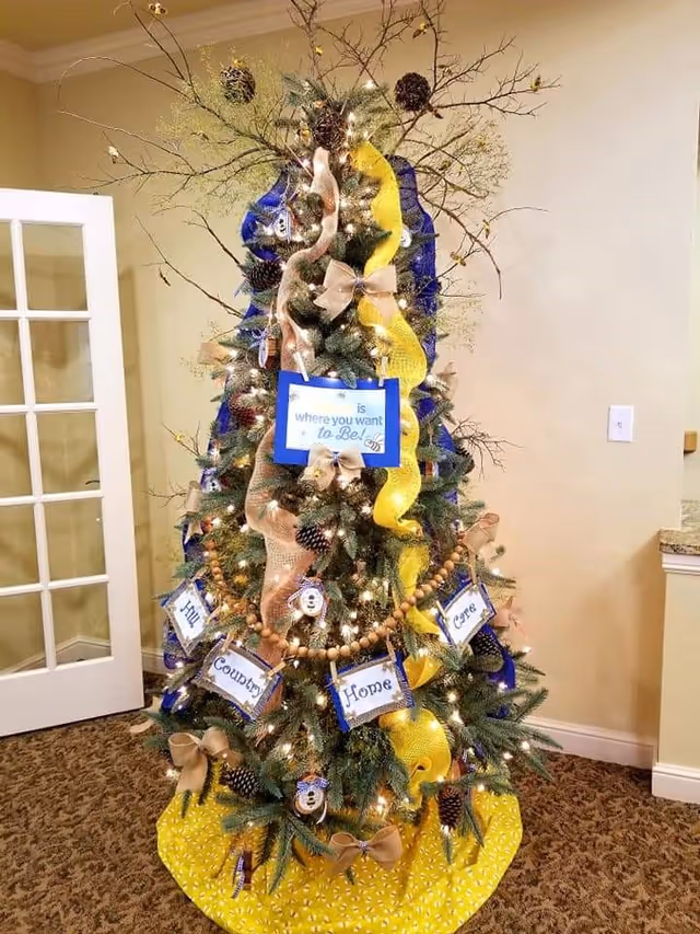 A decorated Christmas tree stands indoors beside a door in a senior living facility.