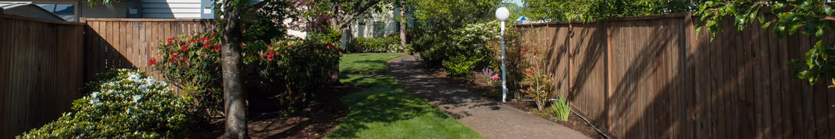 A paved walkway bordered by green grass, flowering bushes, and trees, with wooden fences on both sides and a white lamp post on the right side.