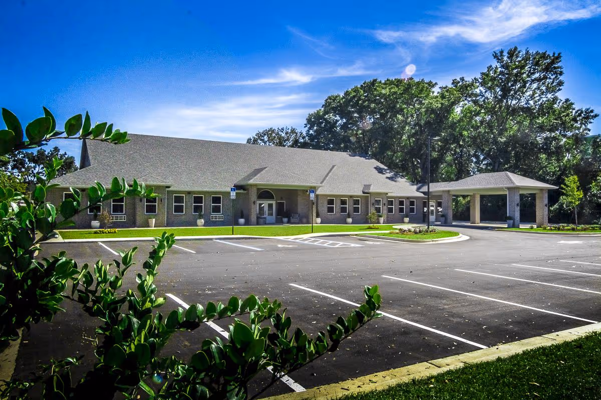 Single-story senior living building with a covered drop-off entrance, empty parking lot, and trees in the background.