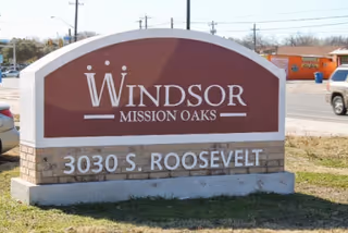 A large outdoor sign for Windsor Mission Oaks with the address 3030 S. Roosevelt displayed below the facility name. The sign is set on a stone base with a maroon and white color scheme. In the background, there is a street with vehicles and some buildings.