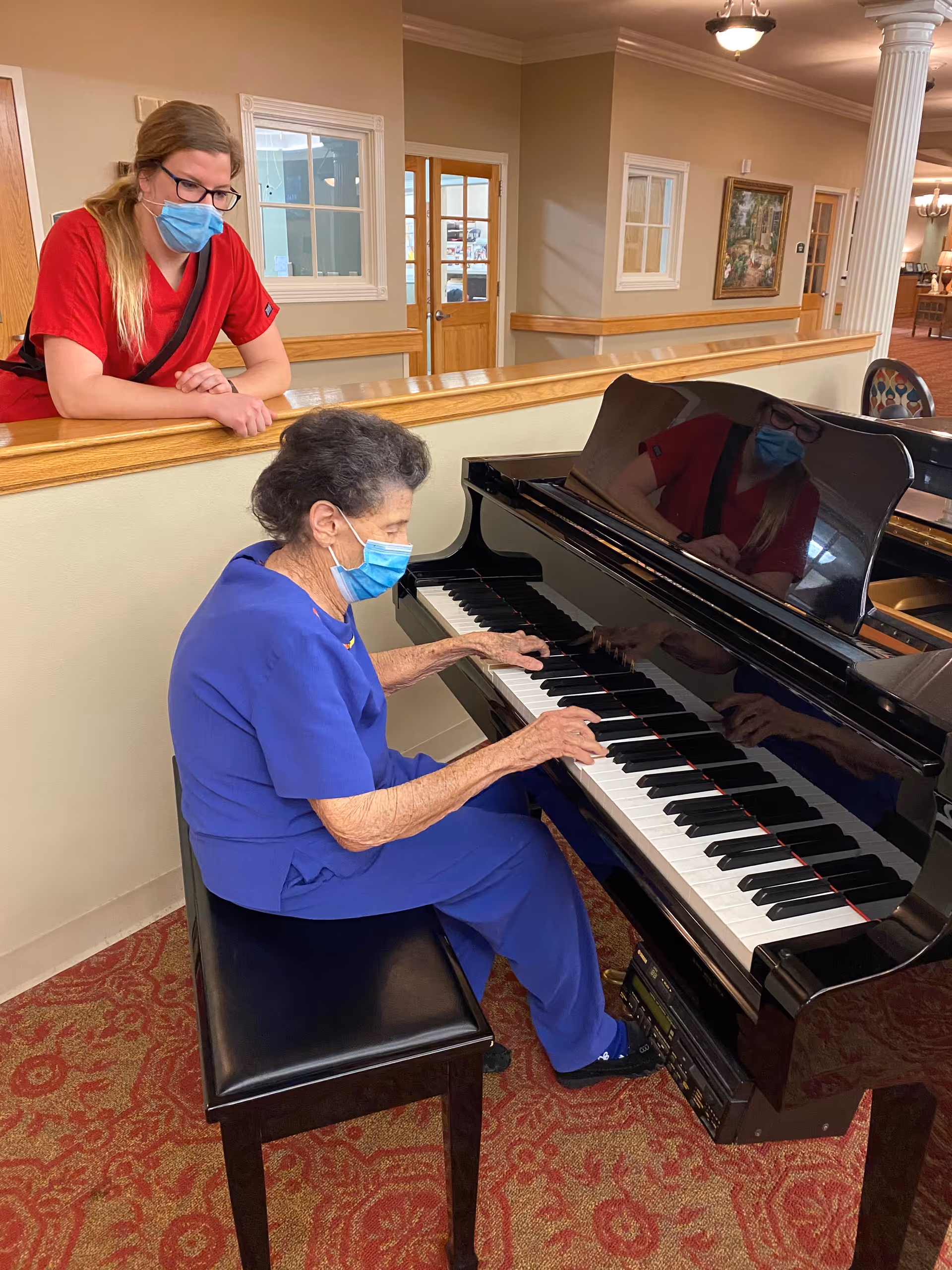 An elderly woman wearing a blue outfit and a face mask is seated at a black grand piano playing music. A younger woman in a red outfit and face mask is leaning on a wooden railing, watching her play. The setting appears to be an indoor common area with beige walls, carpeted floor, and framed artwork on the wall.