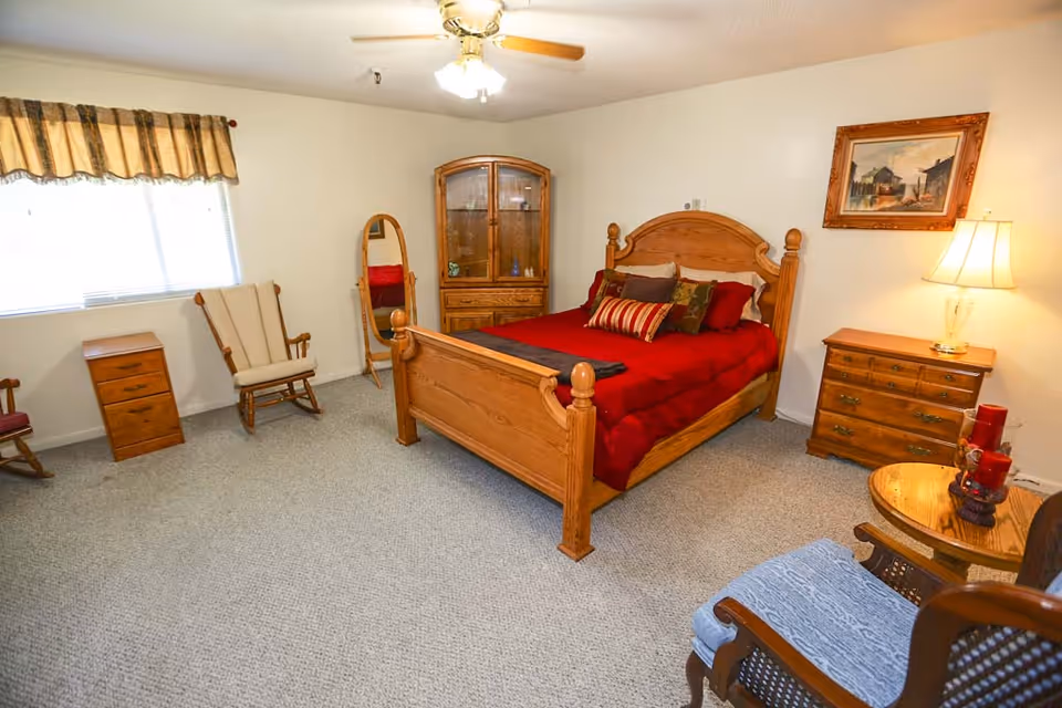 A cozy bedroom with a wooden bed frame featuring a red bedspread and multiple pillows. The room includes wooden furniture such as a dresser with a lamp, a corner cabinet, a small table with red candles, and two chairs. A window with a valance allows natural light to enter, and a framed painting hangs on the wall.