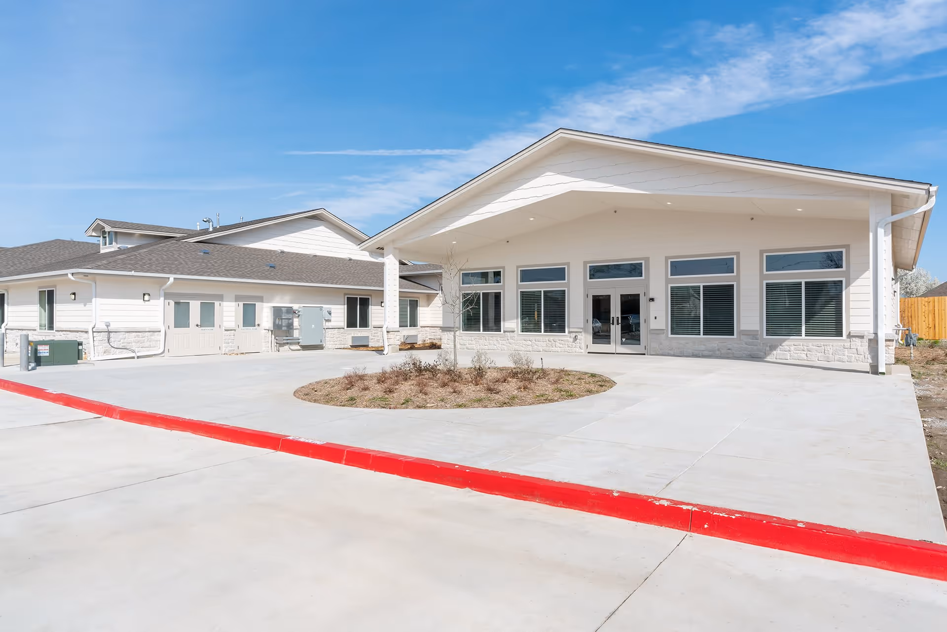 Front exterior of a single-story senior living building with a covered entrance, large windows, and a circular driveway with a red-painted curb.