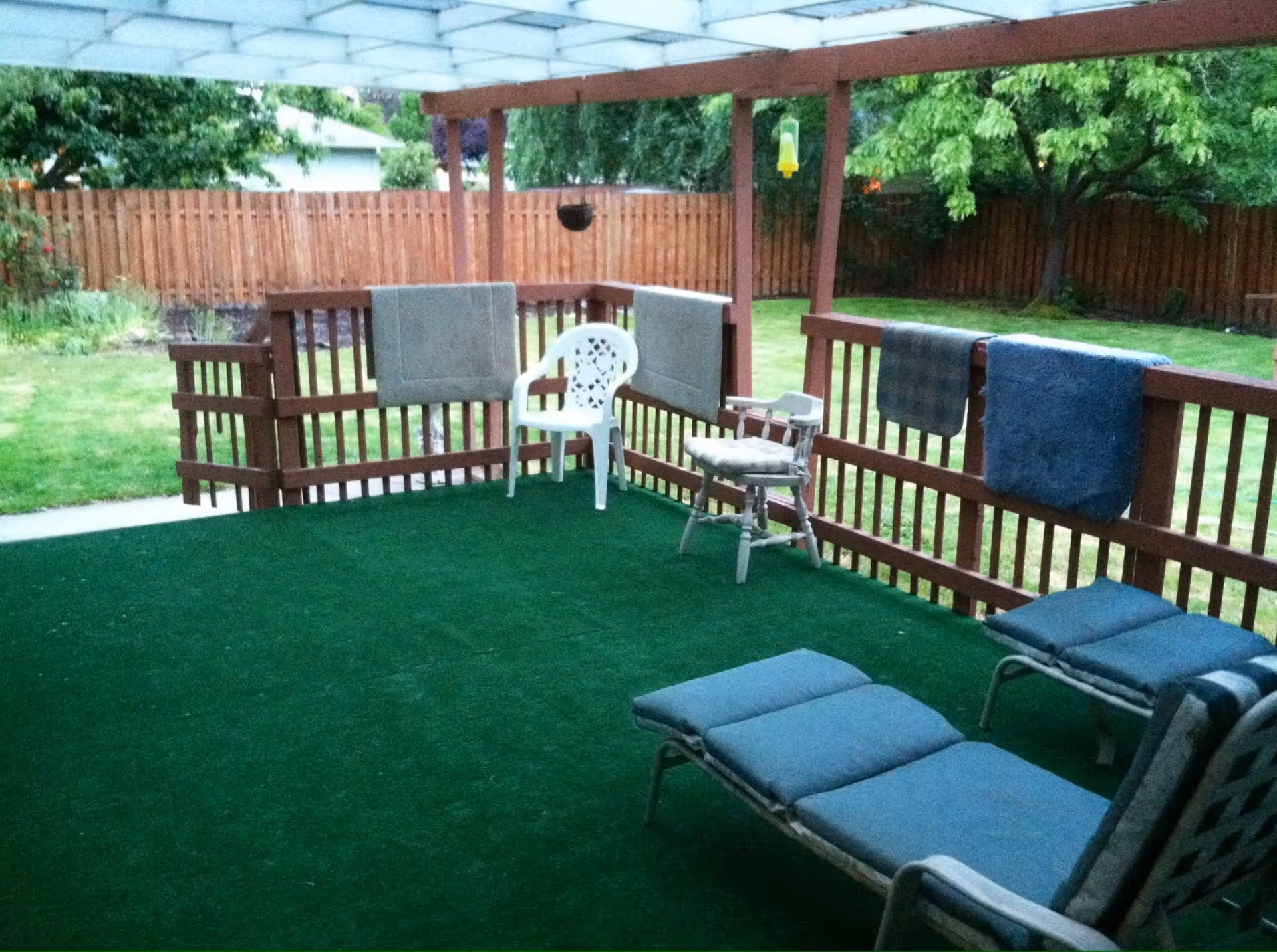 Covered outdoor patio area with green artificial turf flooring, wooden railing with towels hanging on it, two lounge chairs with blue cushions, a white plastic chair, and a wooden chair with a cushion. The patio overlooks a fenced backyard with green grass and trees.