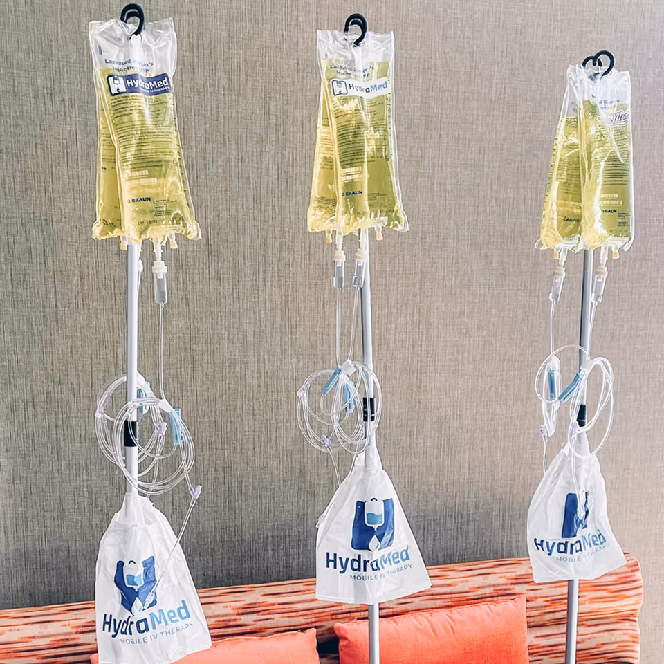 Three HydraMed IV bags on poles stand beside a red couch indoors.