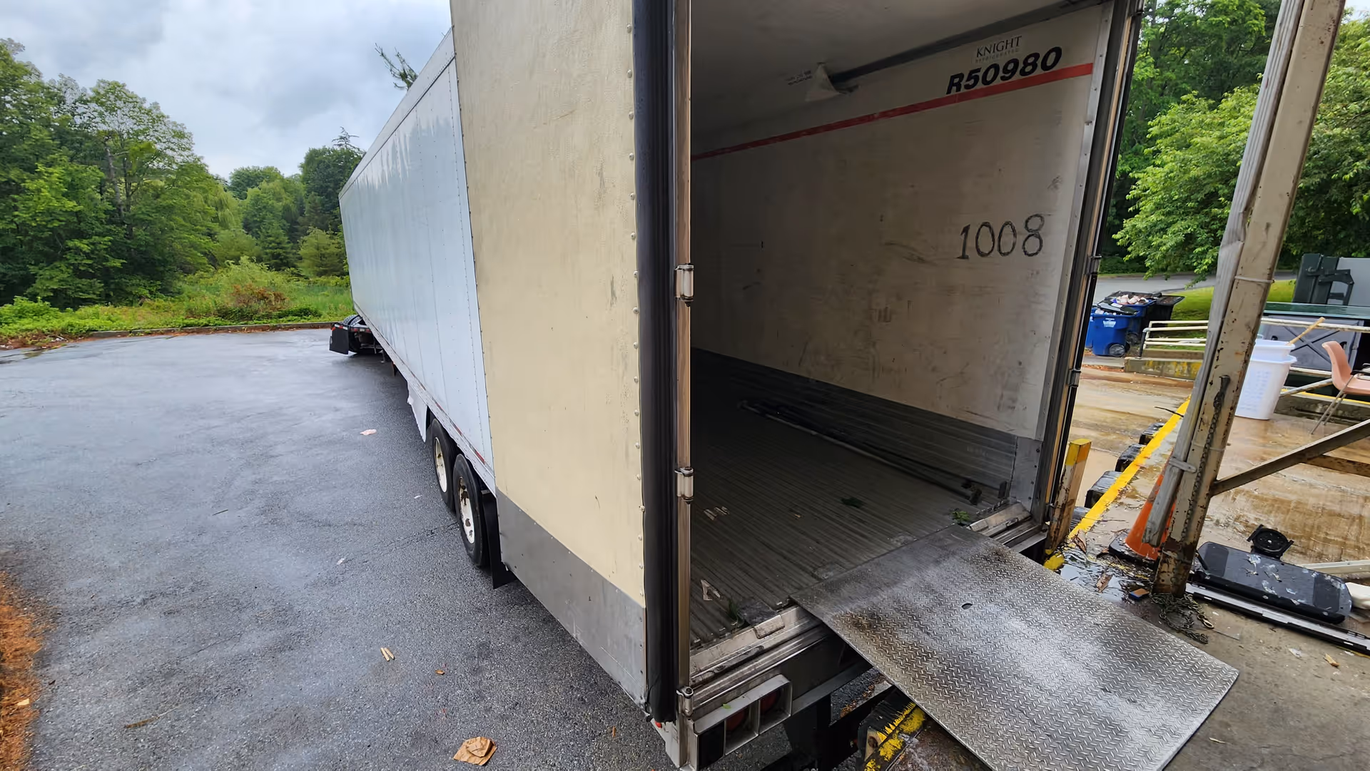 Open trailer at a loading dock with ramp lowered, parked on wet asphalt beside greenery.