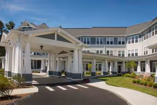 Exterior view of a large, multi-story senior living facility with a covered entrance driveway, landscaped greenery, and a clear blue sky.