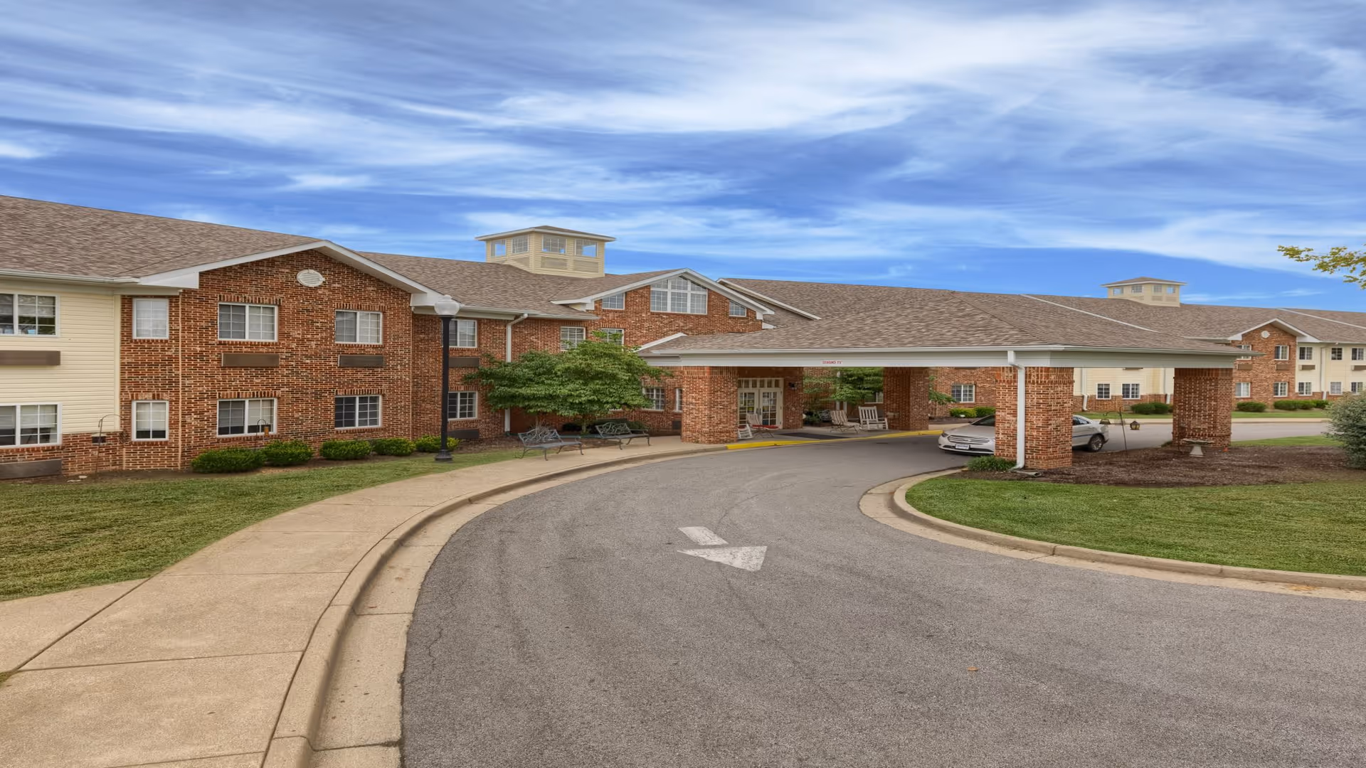Exterior view of McDowell Place of Danville, showing a large brick building with multiple windows and a covered driveway entrance. The sky is partly cloudy, and there is a curved driveway with a white arrow painted on the asphalt. Some greenery and benches are visible near the building.