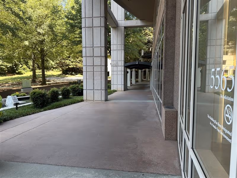 Covered outdoor walkway along a modern building with glass windows and surrounding trees.