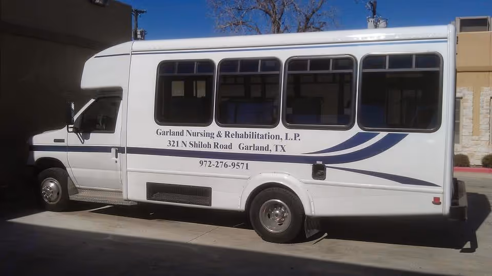 White shuttle bus parked outside a building with the text 'Garland Nursing & Rehabilitation, L.P. 321 N Shiloh Road Garland, TX 972-276-9571' on its side.