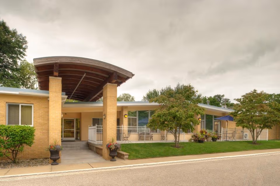 Exterior view of South Haven Nursing & Rehab building entrance with a covered walkway supported by brick pillars, surrounded by green trees and shrubs under a cloudy sky.
