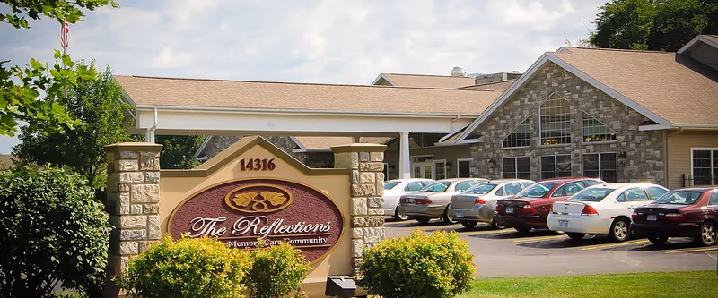 The Reflections Memory Care community sign and the facility's building exterior with a parking lot of cars in front.