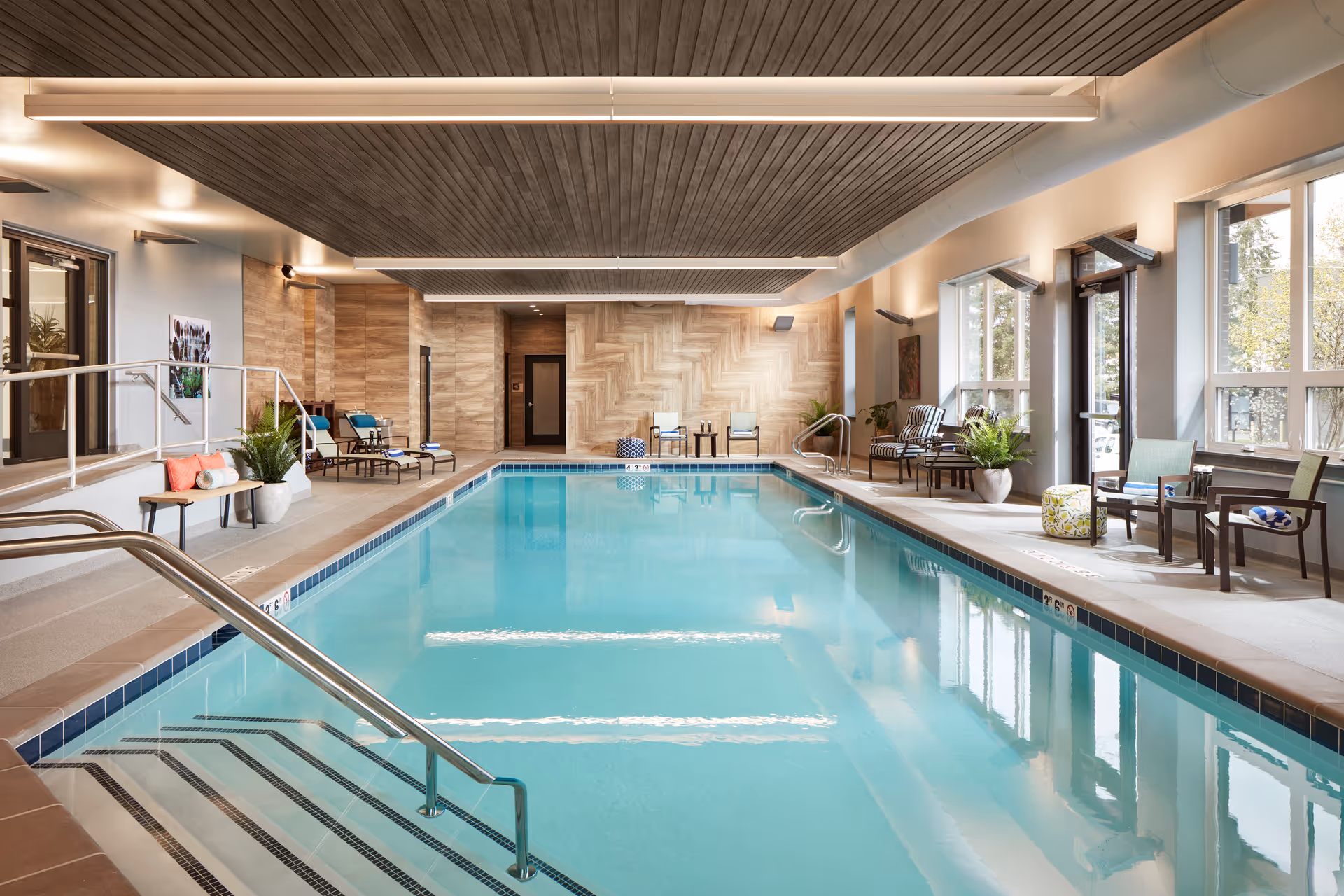 Well-lit indoor swimming pool with lounge chairs, plants, and large windows in a senior living facility.