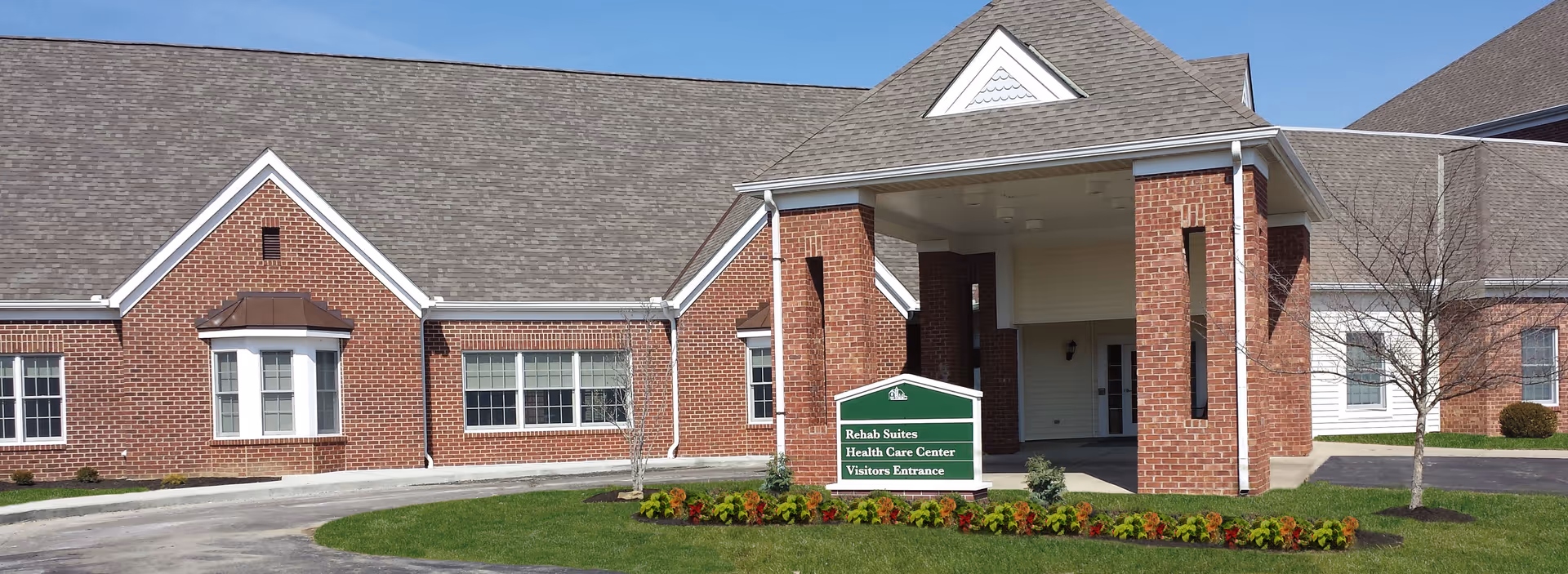 Exterior view of a brick building with a covered entrance and a green sign in front that reads 'Rehab Suites Health Care Center Visitors Entrance'. The building has a gray shingled roof and several windows. There is a curved driveway and a small landscaped area with grass and flowers in front.