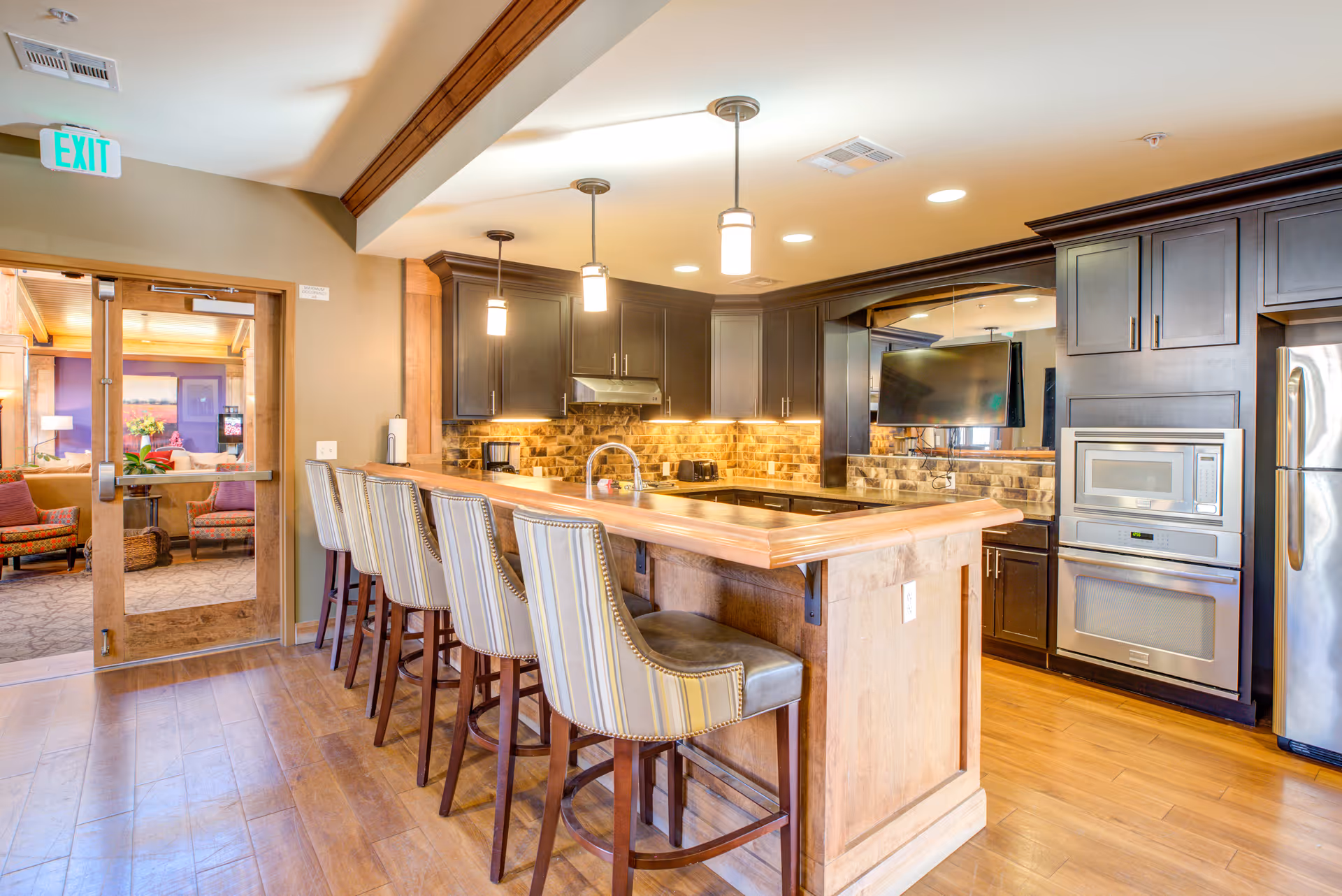 A modern kitchen with a wooden floor and a large wooden island with five striped upholstered bar stools. The kitchen features dark cabinetry, stainless steel appliances including a microwave and refrigerator, a brick-style backsplash, and pendant lights hanging above the island. A glass door leads to a cozy seating area with armchairs and a TV.