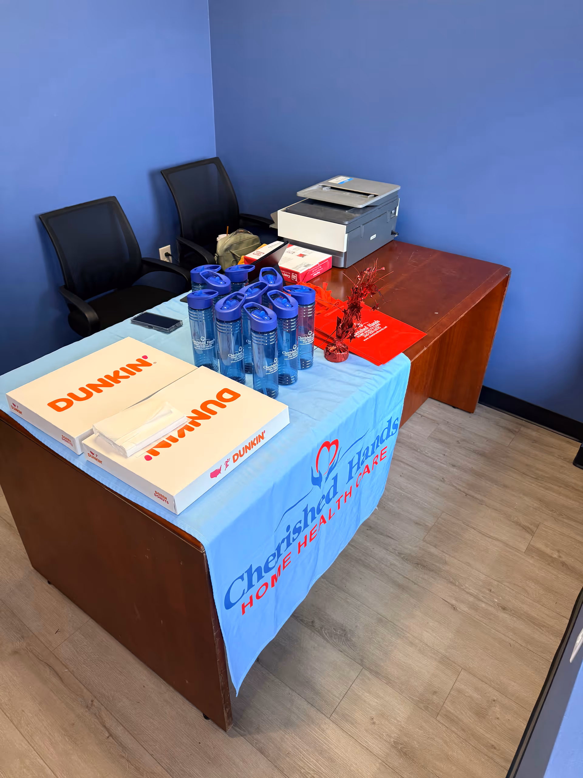 Table in a blue-walled room with Dunkin boxes, water bottles, and a printer.